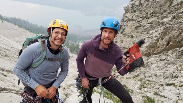 Two men standing high up in a mountain with a lake behind them. 